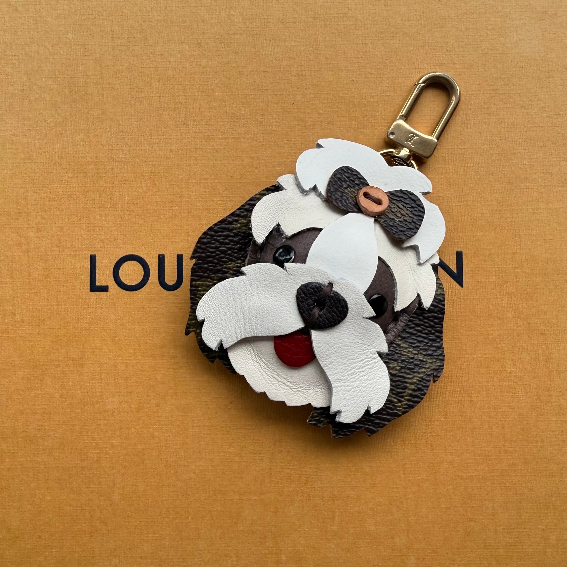 A small dog-shaped keychain bag charm made from layered white and black textured leather, with a gold-tone clasp, photographed on a tan background.
