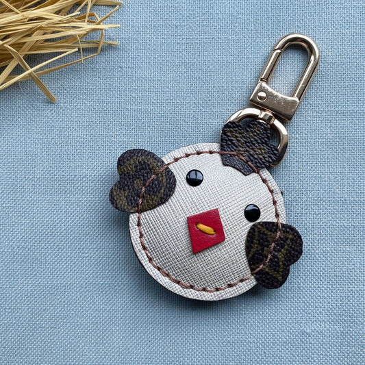 A handmade key chain bag charm featuring a stylized chicken face crafted from textured up-cycled fabrics. The chicken has a light beige textured face, dark brown patterned ears and eyes, a small red stitched nose, and a gold metal keyring clasp.