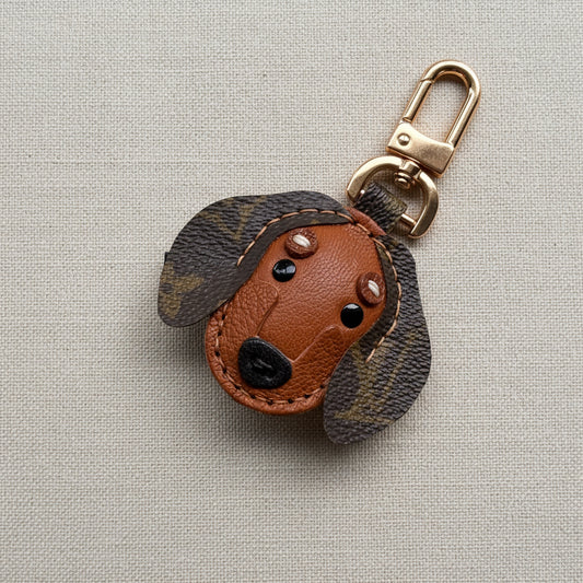 A dachshund-shaped leather keychain bag charm with a brown leather face, dark luxury canvas patterned ears, and a gold metal clasp, displayed on a textured mustard yellow background.